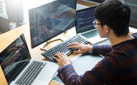 A picture of a person working on the computer and two laptops placed on the left and right sides of his computer.