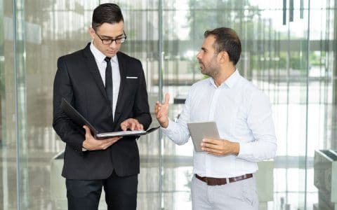 An image shows two persons having a discussion, one person holding a file and the other a tablet in his hand.